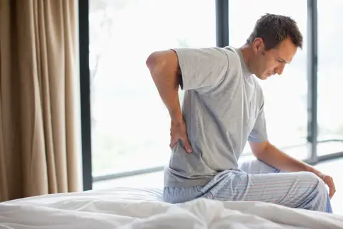 photo of man grabbing his lower back while getting out of bed