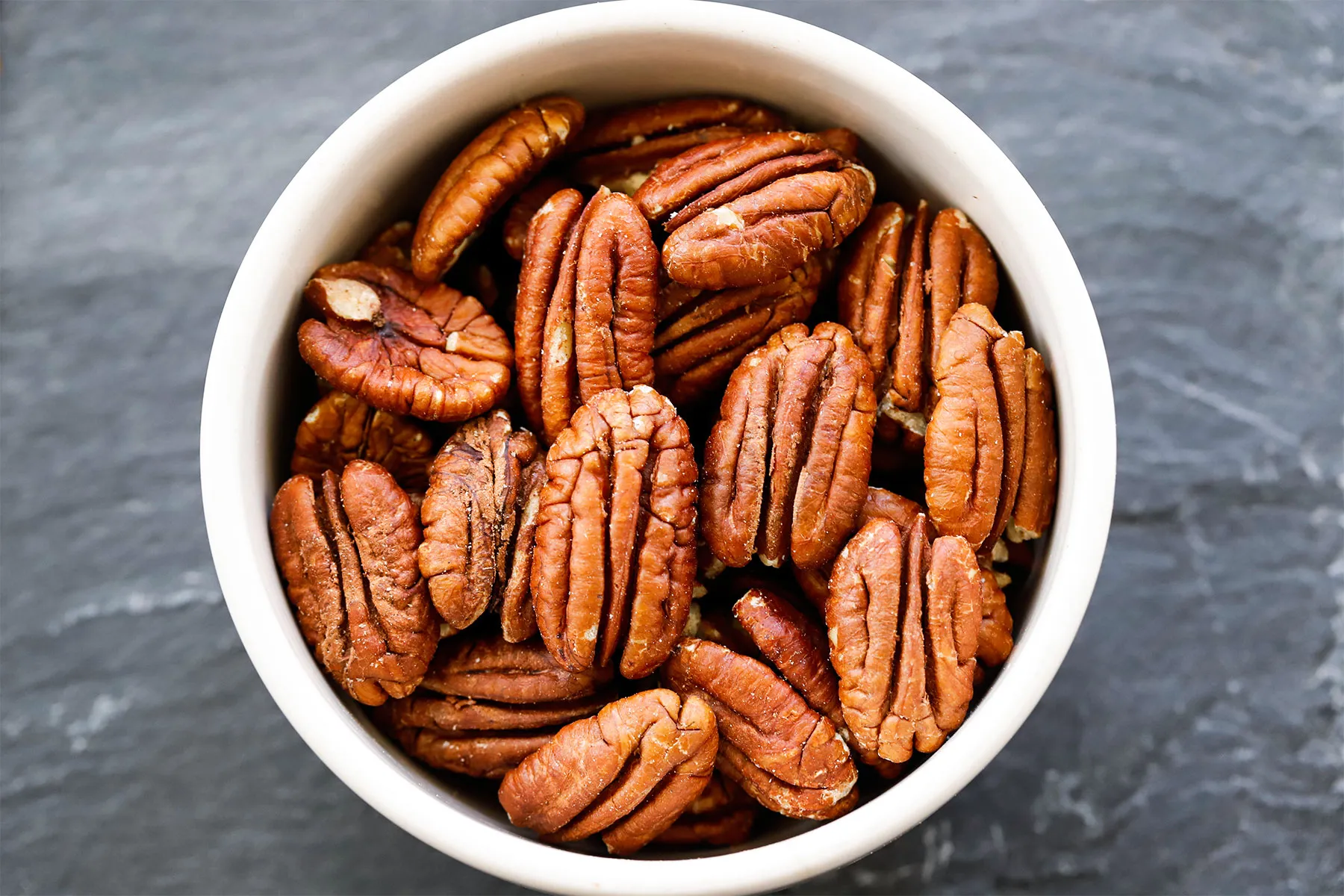 Top-down close-up of a bowl filled with pecan halves on a dark slate-like surface