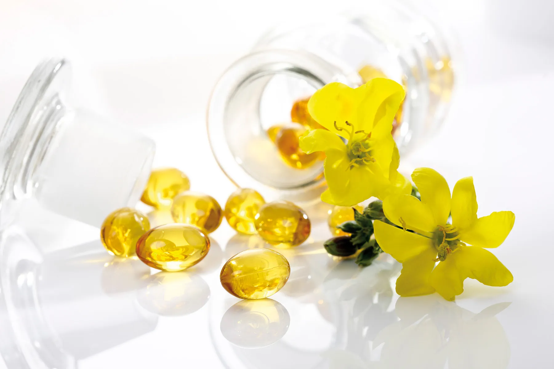 The image presents a still life composition focused on evening primrose oil and its source, the evening primrose flower. A clear glass bottle lies on its side, partially open, with several golden-yellow, translucent softgel capsules scattered around it on a highly reflective white surface. The capsules appear to be made of gelatin and contain a clear, oily liquid. A vibrant yellow evening primrose flower, with four delicate petals and a cluster of stamens, rests near the capsules, its green stem and leaves providing a contrasting color. The flower seems to be freshly picked, with a slight dewiness visible on the petals. The entire scene is brightly lit, creating strong highlights and soft shadows that emphasize the glossy texture of the capsules and the delicate structure of the flower. The white background and reflective surface contribute to a clean, clinical aesthetic, suggesting a focus on health, wellness, or pharmaceutical products. The composition is carefully arranged, drawing the eye from the bottle and capsules to the flower, visually linking the supplement to its natural origin. The overall impression is one of purity, natural ingredients, and a focus on health benefits. The shallow depth of field keeps the focus on the capsules and flower, blurring the background slightly.