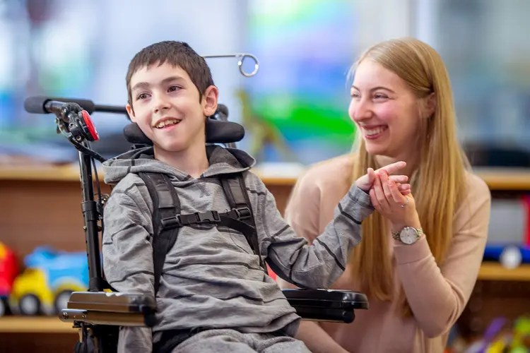 photo of young boy with spinal muscular atrophy sitting in a wheelchair, his teacher hold his left hand.