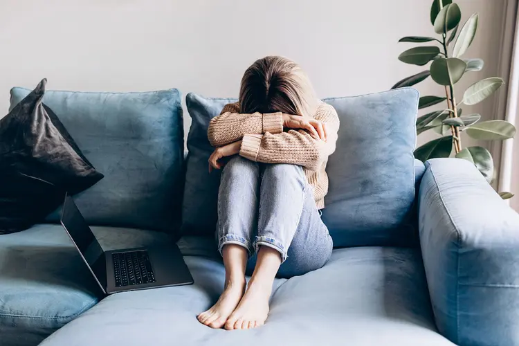 photo of a woman with anhedonia, her head is down on her knees as she sits on a blue couch on the couch with her knees up to her chest