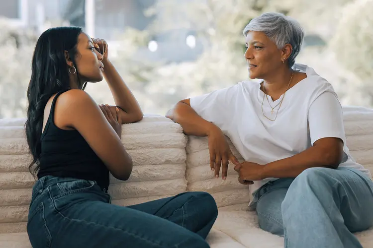 photo of senior mother talking with daughter about her EGFR lung cancer while sitting on a couch accross from each other, an open window behind them shows a blurred-out brightly lit day with trees in the distance