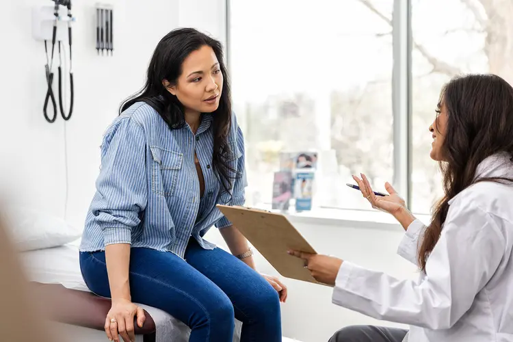 A woman in a blue, quarter-sleeve blouse sitting on exam table and talking about her health with doctor holding a clipboard