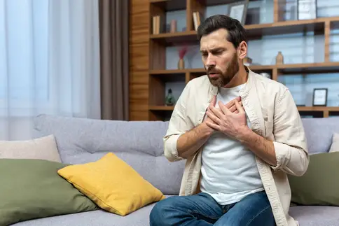 photo of A young man sitting on the sofa at home and holding his chest experiencing shortness of breath 