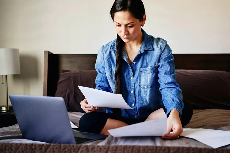 A photo of woman in a denim jacket sitting on a bed with her laptop, surrounded by papers, as she looks at medical bills